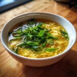 A close-up of a white bowl filled with golden egg drop soup, topped generously with fresh, sliced green onions.