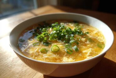A close-up of a white bowl filled with golden yellow egg drop soup, topped generously with fresh green scallions.
