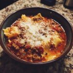 A close-up of a dark bowl filled with rich instant pot lasagna soup, topped with melted cheese and meat sauce.