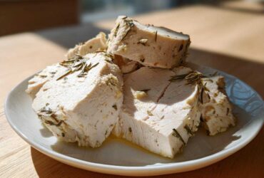 Slices of juicy crockpot turkey breast seasoned with rosemary and garlic, resting on a white plate.