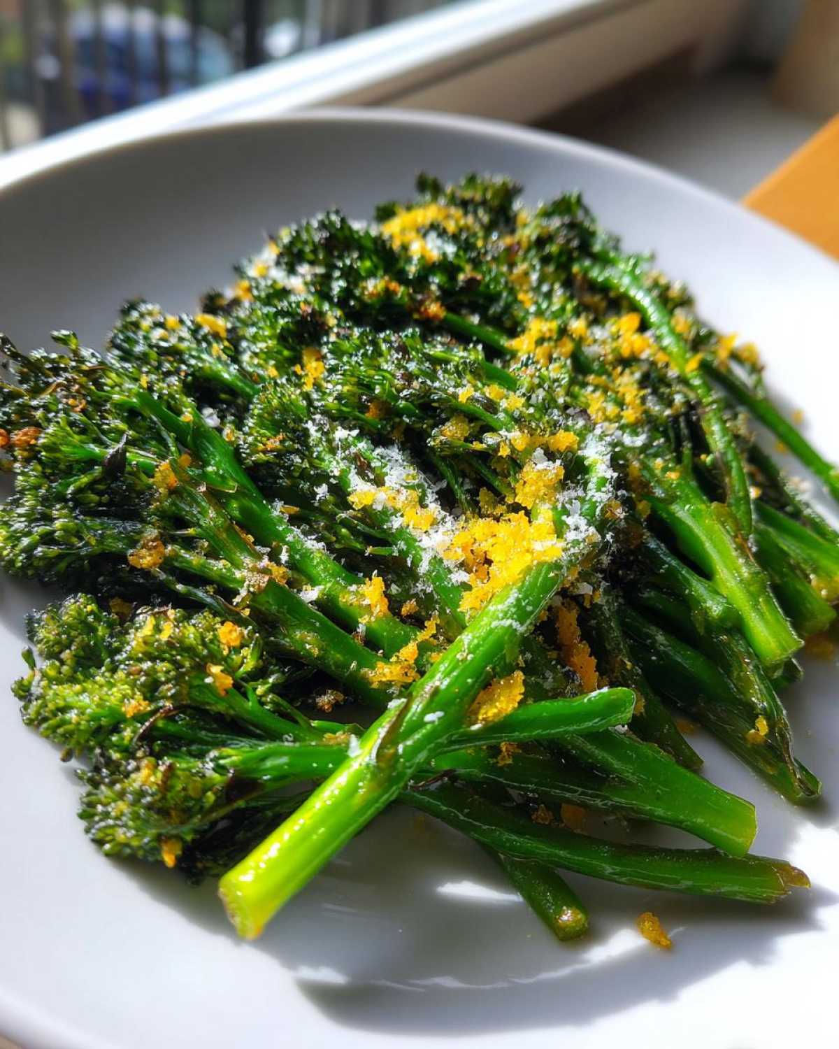 Bright green, cooked broccolini topped with yellow lemon zest and grated cheese on a white plate.