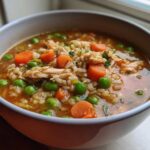 Close-up of a bowl filled with flavorful turkey soup featuring shredded turkey, brown rice, bright green peas, and sliced carrots.