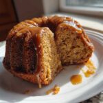 A slice cut from a moist apple bundt cake topped with glossy caramel sauce, served on a white plate.
