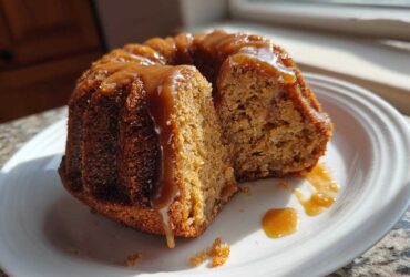 A slice cut from a moist apple bundt cake topped with glossy caramel sauce, served on a white plate.