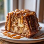 A moist slice of apple dapple cake topped with thick caramel sauce, showing chunks of baked apple inside.