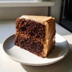 A rich, dark slice of gingerbread cake topped with thick, light brown frosting, sitting on a white plate.