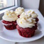 Four vibrant red velvet cupcakes topped with swirls of rich cream cheese frosting, sitting on a white plate.