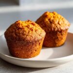 Close-up of two golden brown, moist sweet potato muffins sitting on a light-colored plate.