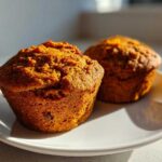 Two freshly baked, golden-brown sweet potato muffins resting on a small white plate, highlighted by sunlight.