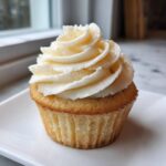 Close-up of a single, perfectly baked vanilla cupcake topped with a tall swirl of creamy vanilla frosting.