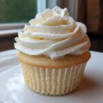 Close-up of a single, perfectly baked vanilla cupcake topped with a tall swirl of white vanilla buttercream frosting.