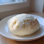 A freshly made, smooth ball of white fufu served on a small white plate, ready to eat.