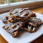 A stack of rich peanut butter pretzel bark pieces drizzled with white chocolate on a white plate.