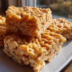 Close-up of stacked, glossy peanut butter rice krispie treats squares on a white plate.