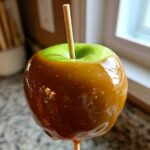 Close-up of a shiny, homemade caramel apples dipped over a bright green Granny Smith apple with a wooden stick inserted.