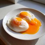 Two perfectly cooked poached eggs on a white plate, showing bright orange runny yolks spilling out.