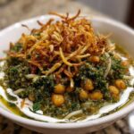 A close-up of a bowl of rich, green persian noodle soup topped with chickpeas, fried onions, and a swirl of white yogurt.
