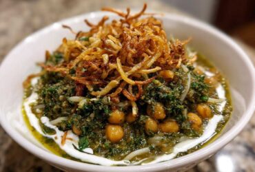 A close-up of a bowl of rich, green persian noodle soup topped with chickpeas, fried onions, and a swirl of white yogurt.