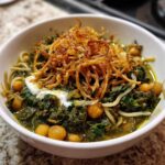 A close-up of a bowl of rich, green persian noodle soup (Ash Reshteh) garnished with crispy fried onions and a dollop of yogurt.