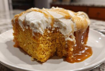 A moist slice of orange pumpkin poke cake topped with white frosting and drizzled with caramel sauce.
