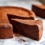 A decadent slice of flourless chocolate torte dusted heavily with cocoa powder, sitting next to the main cake.