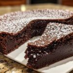 A decadent flourless chocolate torte dusted with powdered sugar, with one slice cut and resting beside the main cake.