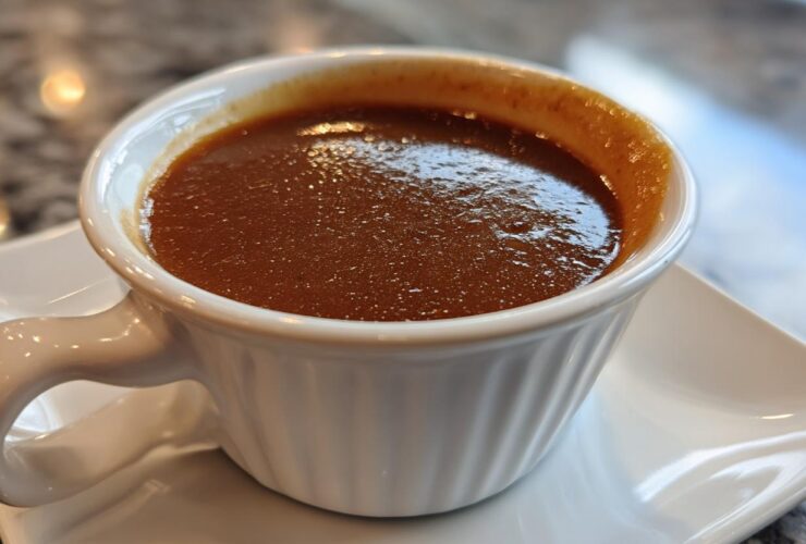Close-up of rich, dark homemade brown gravy served in a small, white, handled ramekin on a matching saucer.