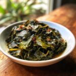 A close-up of a white bowl filled with rich, savory collard greens simmered in broth, sitting on a wooden surface.