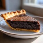 A close-up slice of rich, dark chocolate chess pie with a flaky crust, sitting on a white plate.