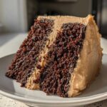 A close-up slice of dark, rich gingerbread cake layered and covered with light brown frosting.