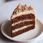 A tall slice of dark chocolate gingerbread latte cake with layers of cream filling and spiced frosting peaks.