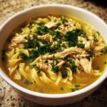 Close-up of a white bowl filled with rich broth, egg noodles, shredded chicken, and fresh parsley, representing slow cooker chicken noodle soup.