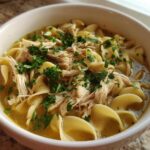 Close-up of a white bowl filled with rich broth, wide egg noodles, shredded chicken, and fresh parsley, showcasing the slow cooker chicken noodle soup.