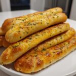 A stack of freshly baked, soft garlic breadsticks topped with melted cheese and chopped parsley on a white plate.