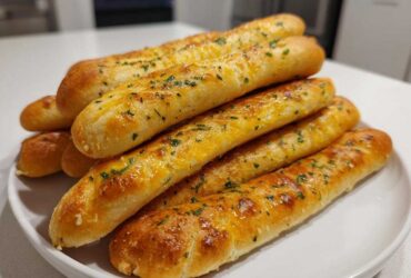 A stack of freshly baked, soft garlic breadsticks topped with melted cheese and chopped parsley on a white plate.