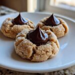 Three soft peanut butter blossom cookies rolled in sugar, each topped with a chocolate kiss, displayed on a white plate.