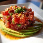 A mound of bright red spicy tuna tartare served over fanned slices of fresh avocado on a white plate.