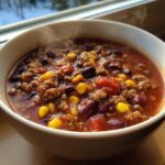 A steaming white bowl filled with hearty crockpot cowboy soup featuring ground beef, kidney beans, corn, and tomatoes.