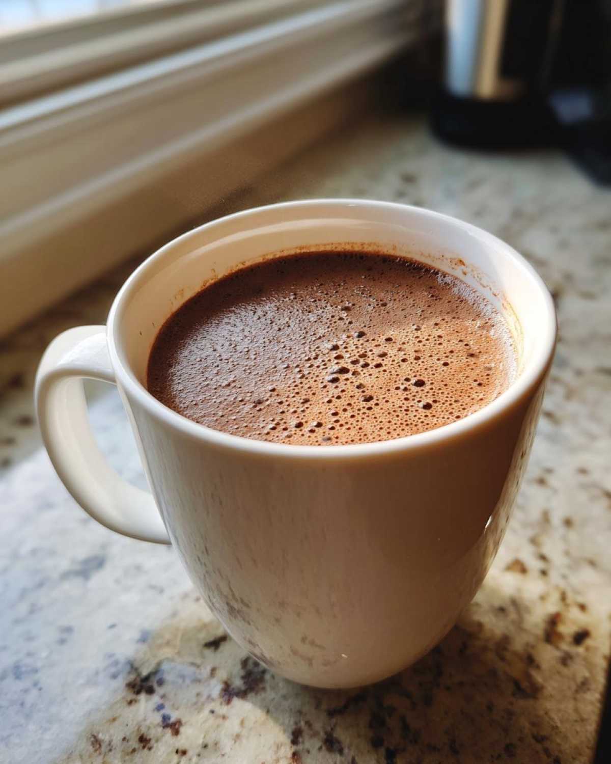 Close-up of a steaming white mug filled with rich, foamy hot cocoa mix beverage on a granite countertop.