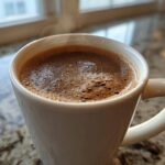 Close-up of a steaming white mug filled with rich, frothy hot cocoa made from hot cocoa mix, sitting on a granite counter.
