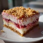 A close-up, tempting slice of strawberry crunch cheesecake showing layers of crust, cream, and strawberry filling, topped with golden crumble.
