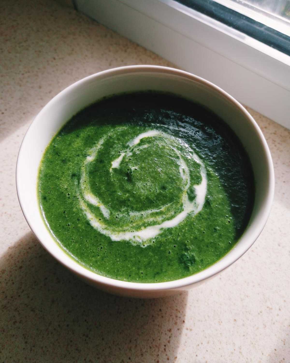 A close-up overhead view of a bowl of bright green spinach soup, garnished with a swirl of white cream.