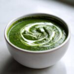 Close-up of a bowl of rich green spinach soup topped with a swirl of white cream.