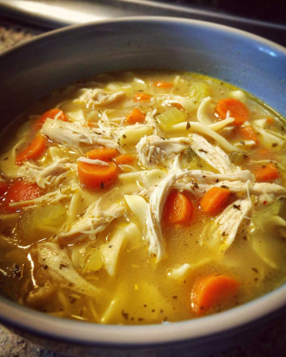 Close-up of a blue bowl filled with rich broth, shredded chicken, wide noodles, and bright orange carrots in chicken noodle soup.