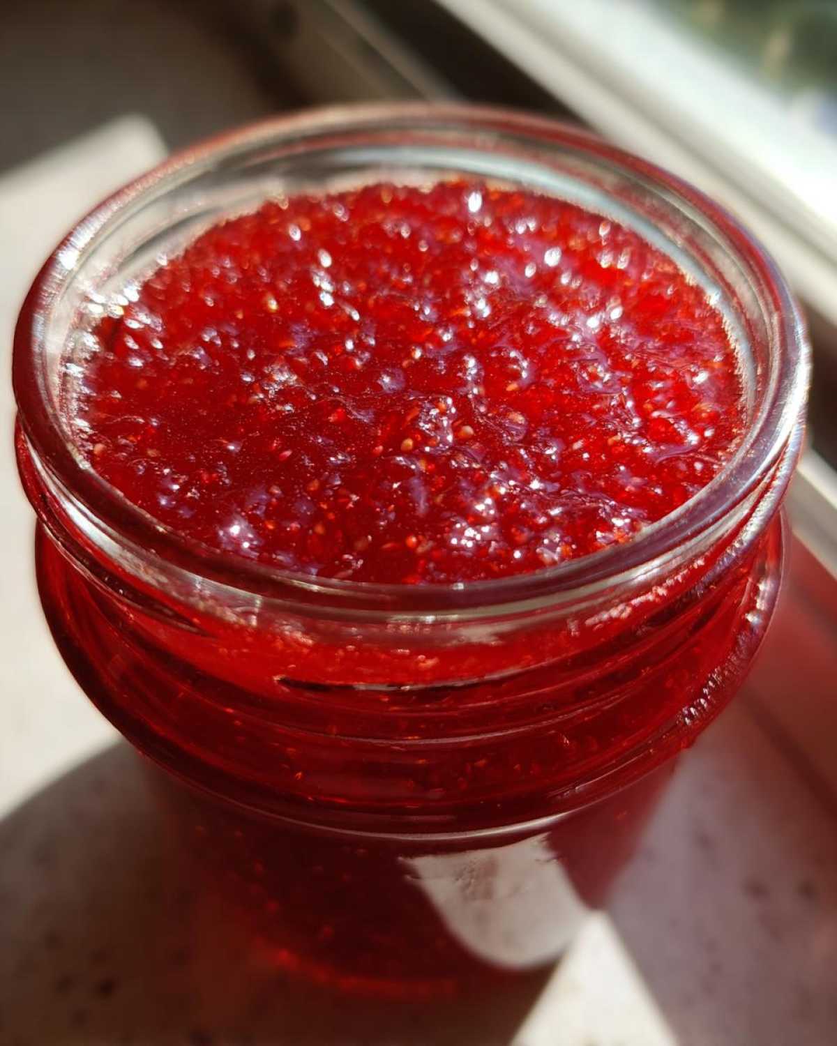 Close-up of bright red, glistening homemade raspberry jam filling a glass jar, showing visible seeds.