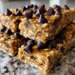 A stack of homemade no-bake protein bars topped generously with dark chocolate chips, sitting on a granite counter.