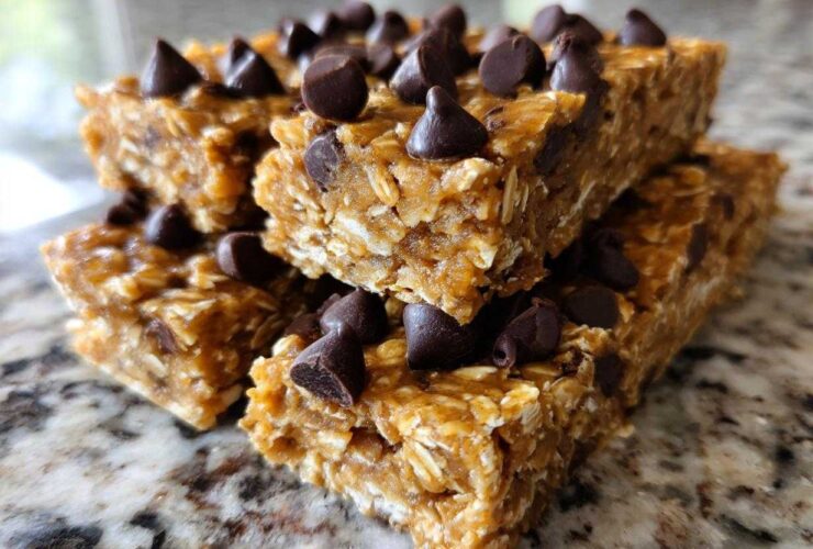 A stack of homemade no-bake protein bars topped generously with dark chocolate chips, sitting on a granite counter.