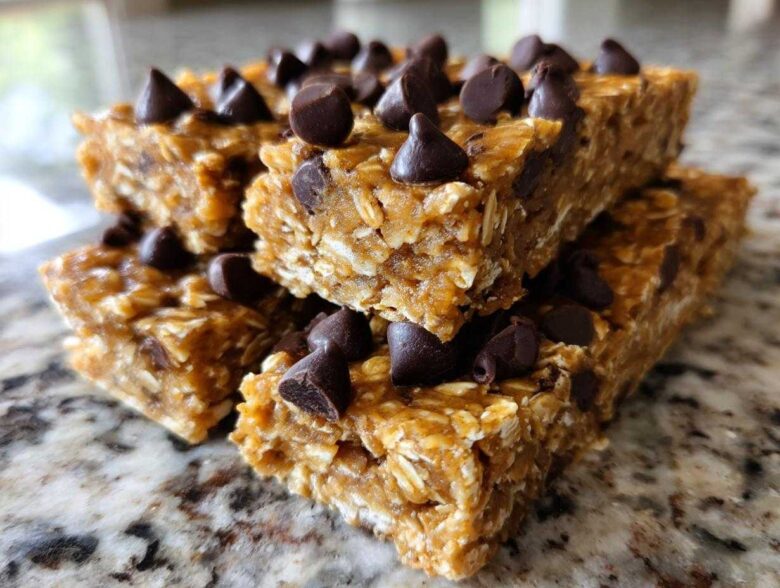 A stack of homemade no-bake protein bars topped generously with dark chocolate chips, sitting on a granite counter.