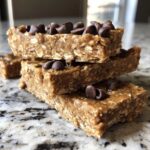 A stack of three homemade, no-bake protein bars topped with mini chocolate chips on a granite counter.