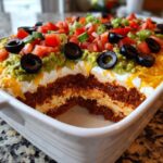A white dish showing a cross-section of a delicious 7 layer dip topped with guacamole, tomatoes, olives, and cheese.